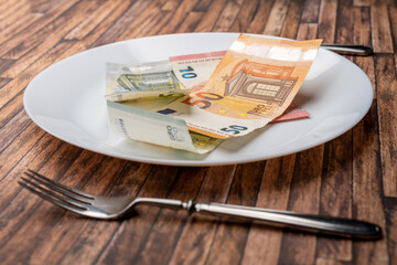 Euro banknotes lying on a dinner plate with cutlery on the table. Symbolic image for eating money notes. Food is getting more expensive due to inflation effects. Hunger caused by a crisis.