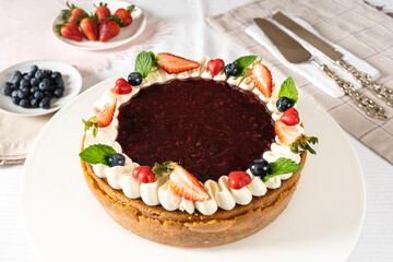 red fruit cheesecake, cream, garnished with pieces of fresh fruit strawberries, blueberry, cherry and mint on white table with cutlery and fruits