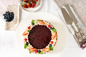 red fruit cheesecake, cream, garnished with pieces of fresh fruit strawberries, blueberry, cherry and mint on white table with cutlery and fruits