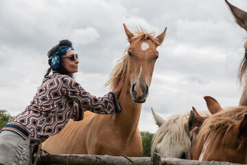 A beautiful girl in glasses and with a bandana on her head strokes the horses in the meadow. Blurred background.