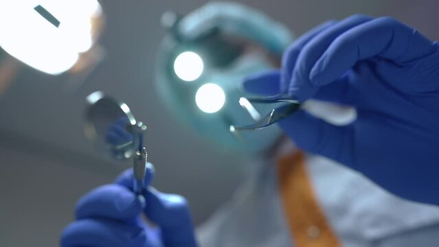 Close-up dental mirror and tweezers in hands of blurred unrecognizable doctor at background. Patient point of view of professional expert dentist with tools in hospital indoors. Slow motion