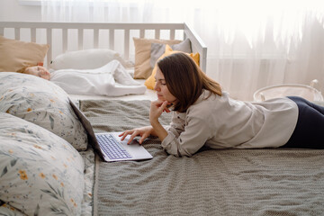 A young mother freelancer working on laptop laying on the bed during baby's daytime sleeping time. Mom woman working from home remotely.