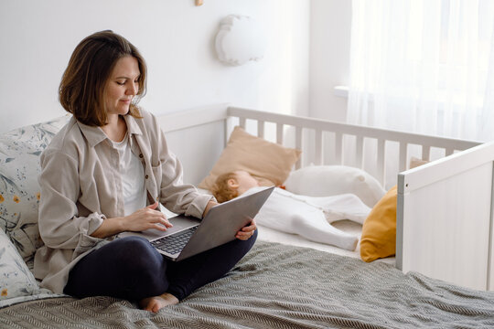Mom freelancer. A young mother works on laptop during quiet time while her baby toddler sleeps on the crib. A woman working home remotely.