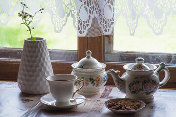 a tea set and a vase with daisies on the table in front of a wooden window of a rural house