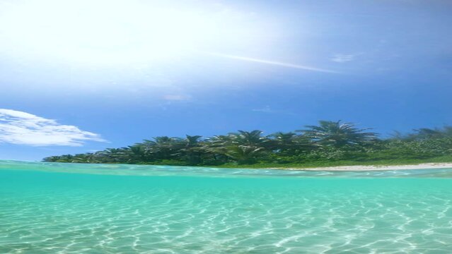 VERTICAL SLOW MOTION, HALF UNDERWATER, LENS FLARE: Hot tropical sunrays shine on picturesque tropical sandy beach. Beautiful small crystal clear ocean waves roll towards the idyllic exotic island.