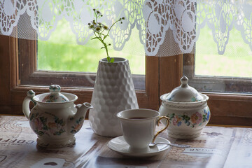 a tea set and a vase with daisies on the table in front of a wooden window of a rural house