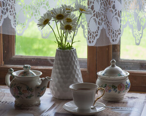 a tea set and a vase with daisies on the table in front of a wooden window of a rural house