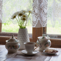 a tea set and a vase with daisies on the table in front of a wooden window of a rural house