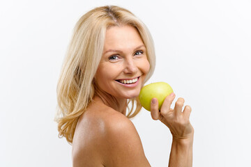 Beautiful playful middle-aged topless woman holding fresh green apple in hand, looking at camera and smiles with toothy smile, dental care concept