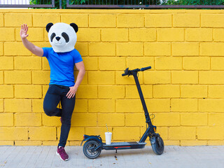Unrecognizable man with panda bear mask waving next to an electric scooter leaning on a yellow wall.