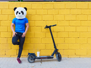 Man with panda bear mask next to an electric scooter leaning on a yellow wall.