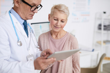 Fototapeta premium Senior practitioner with stethoscope around neck standing in clinic room and answering patients questions according to test results on tablet