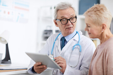 Serious wrinkled senior doctor in lab coat sitting at table and using digital tablet with online patients records while giving recommendations to mature woman