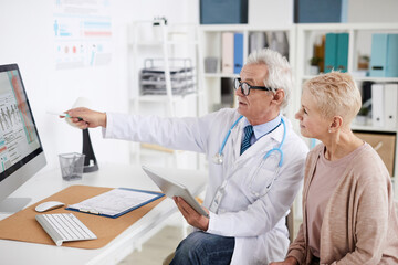 Confident senior male practitioner with gray hair sitting at table and pointing at computer screen while consulting mature female patient in clinic