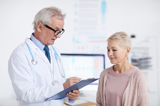 Professional Gray-haired Doctor In Glasses Standing In Clinic Room And Making Notes In Clipboard While Talking To Senior Patient