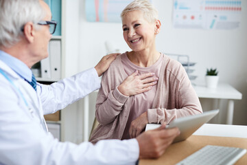 Fototapeta premium High-qualified senior doctor in lab coat sitting at table and using tablet while reporting good tests results to senior patient