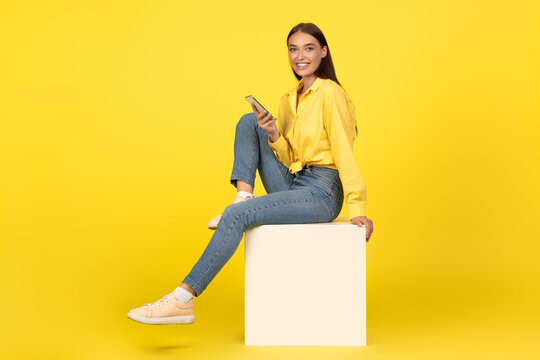 Lady Using Cellphone Smiling To Camera Sitting Over Yellow Background