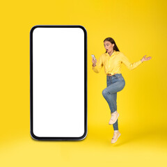 Shocked Woman Holding Phone Posing Near Huge Smartphone, Yellow Background