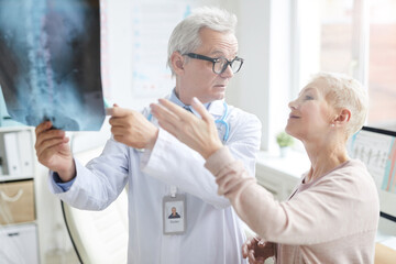 Obraz premium Attractive blond-haired mature lady pointing at core x-ray image while discussing her pain with doctor
