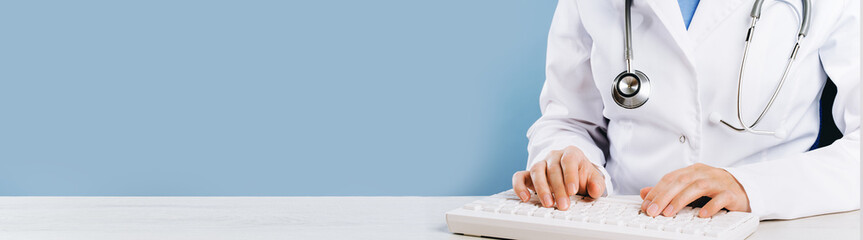Doctor typing on computer keyboard in office. Physician in uniform uses computer at desk. Professional diagnosis and treatment tele medicine. Professional healthcare wide banner - obrazy, fototapety, plakaty