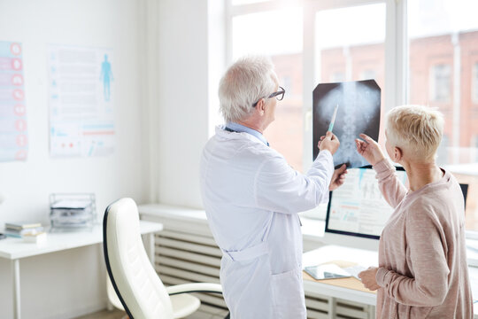 Rear view of gray-haired doctor in lab coat standing against window and watching x-ray image of spinal cord with female patient
