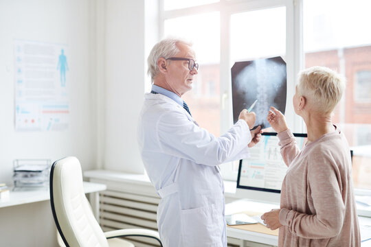 Serious senior male radiologist in white coat standing in own office and showing x-ray image of spine to mature patient