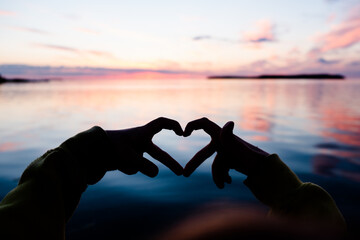 Heart shape hand gesture made with fingers against colorful out of focus sunset sky. Empty copy...