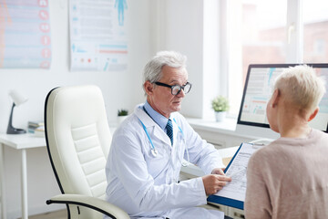 Obraz premium Serious elderly doctor in lab coat and glasses sitting at desk with computer and asking patient to fill form