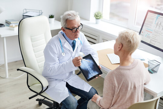 Serious aged doctor in white coat sitting at table and using tablet while showing online x-ray image of spine to patient