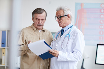 Serious senior doctor in lab coat and glasses holding clipboard with medical papers and watching test results with aged patient © Mediaphotos