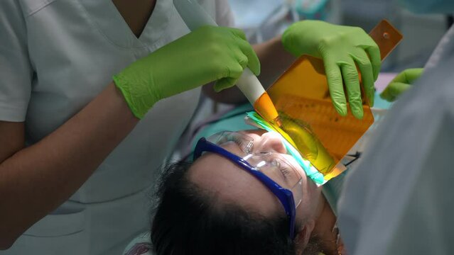 High angle view of Caucasian woman in dental chair with unrecognizable doctor and nurse using UV light for polymerization. Professional expert dentist and assistant treating patient teeth. Slow motion