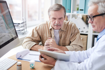 Serious male doctor sitting at desk with computer and showing medical record on tablet to aged patient