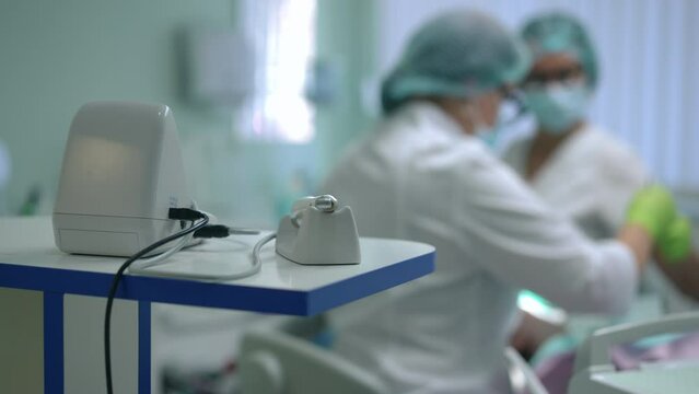 Close-up dental curing light with blurred doctor and nurse treating patient in dental chair at background. Expert professional Caucasian dentist and assistant working with Caucasian woman indoors