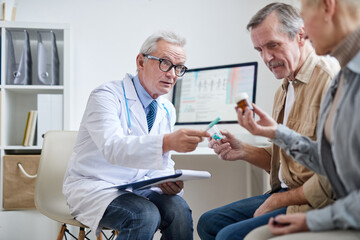 Senior gray-haired doctor with stethoscope around neck pointing at pill bottle in hand of patient and explaining how to take it to patients