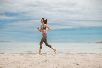 Fototapeta premium Young beautiful sportive woman in headphones running over seaside on morning at beach