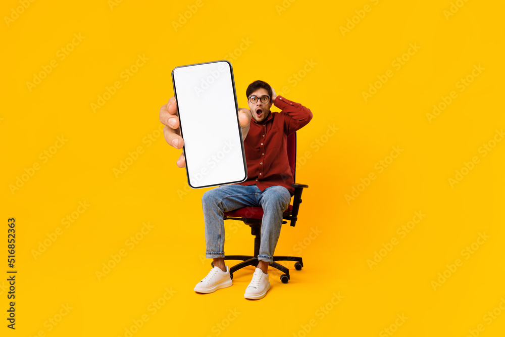 Wall mural Shocked young man sitting in chair and showing big blank smartphone