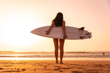 Fotobehang Portrait, a surfer woman on a beach at sunset with the surfboard looking at the sea © unai