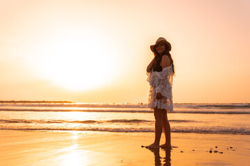Portrait of a woman at sunset in a white dress with a hat walking by the sea at low tide
