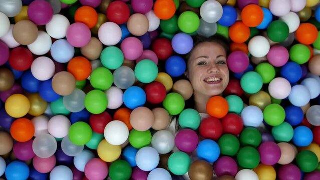 Beautiful girl lies in a pool full of colorful plastic balls. Place for text, cheerful video with girl. Smiling caucasian woman opens her eyes and looks at the camera while among many small balls.