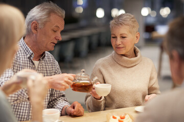 Handsome senior man in casual shirt pouring tea to ladys cup while they having tea party with friends at home