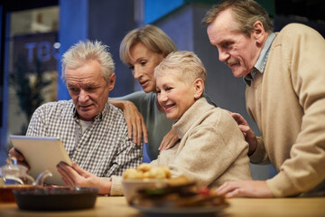 Group of positive senior people sitting at table and using digital tablet while enjoying funny video