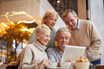 Fototapeta na wymiar Group of positive senior friends in casual clothing using tablet while surfing websites in cafe with wifi