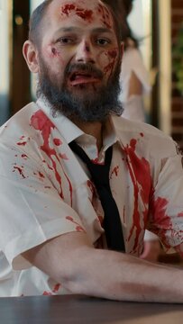 Vertical video: Ugly dirty office zombie sitting at desk while working on modern laptop. Frightening creepy monster with deep and bloody wounds and scars using portable computer for work in company