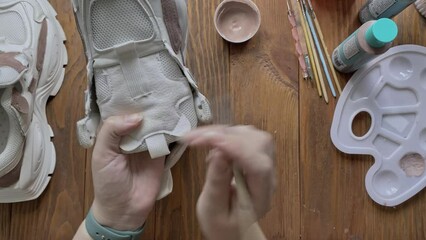 Top view of hands paint white boots with brush on wooden table. Palette with brown acrylic and other tools on desktop. Hobbies, creativity and handmade work at home. Designer's workshop.