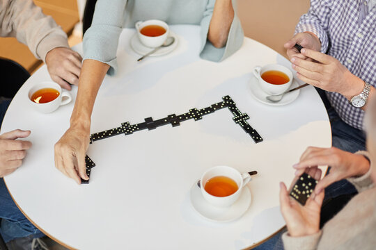 Group of unrecognizable seniors sitting at round table with tea cups and wasting time with domino game