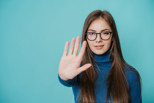 Stop! Stay away! A young European girl in a dark blue jumper and pants put her hand in front of her in a stopping gesture, standing on a turquoise background..Student trying to avoid bullying.