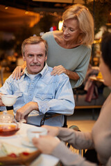 Smiling affectionate blond-haired wife embracing husband in wheelchair while he drinking tea and talking to friends at gathering