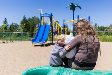 Sweet moment captured from the back of a mother sitting on a playground during a bright sunny day,...