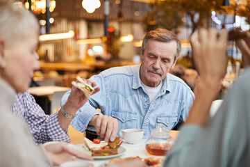 Fototapeta na wymiar Content handsome senior man with mustache sitting at table and eating sandwich while having brunch with friends