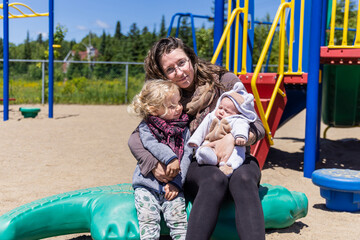 Front portrait of happy smiling mother cradling two children at playground on sunny day. Three year old boy looks at newborn sister. With copy space.